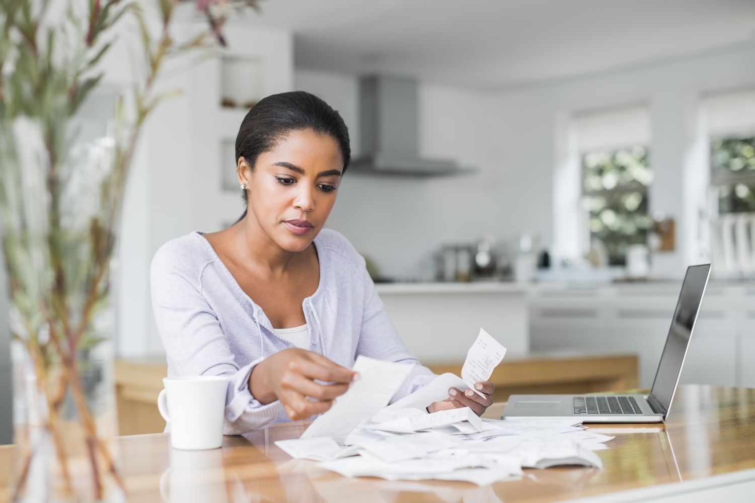 A person sitting at a kitchen table paying bills on a laptop with a mug of coffee nearby